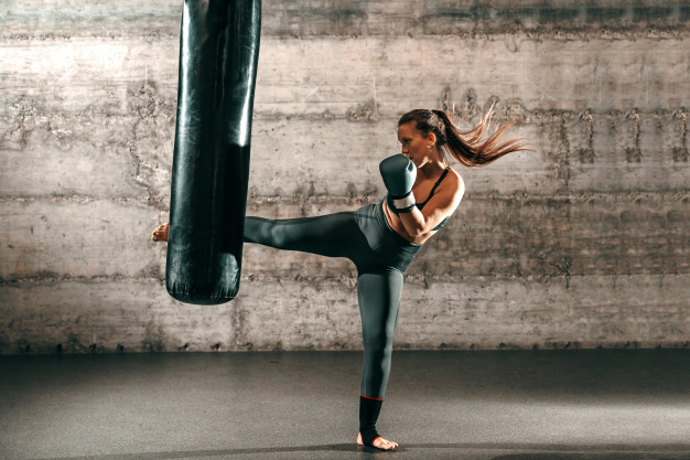 dedicated-strong-brunette-with-ponytail-sportswear-bare-foot-with-boxing-gloves-kicking-sack-gym_232070-5906